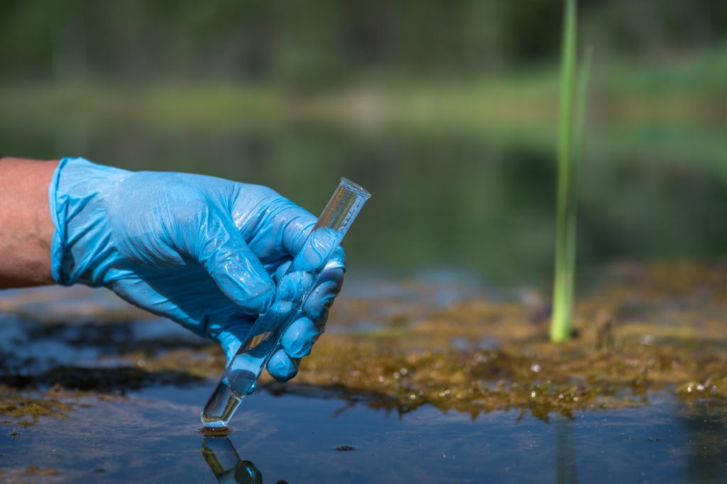 The hand of a working man in a glove holds a test tube with a water sample in close-up against the background of a natural landscape.